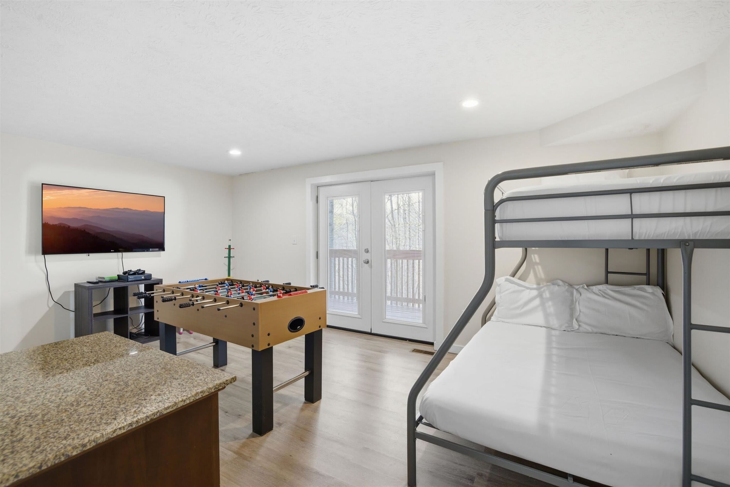 Una habitación con mesa de fútbol, cama con estructura de metal, televisor en la pared y puerta con paneles de vidrio.