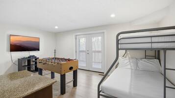 Una habitación con mesa de fútbol, cama con estructura de metal, televisor en la pared y puerta con paneles de vidrio.