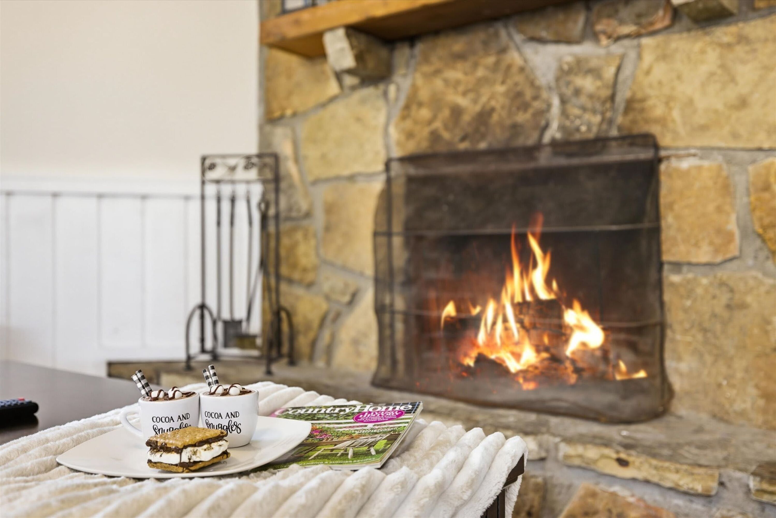 Une pièce chaleureuse avec une cheminée en pierre, une table avec deux tasses et un magazine, et une couverture sur le sol.