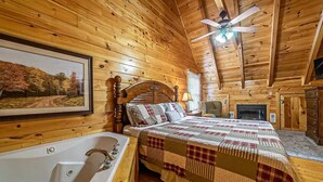 Un dormitorio tipo cabaña de troncos con una cama grande, techo de madera, ventilador de techo, chimenea y un cuadro con una imagen de un bosque.