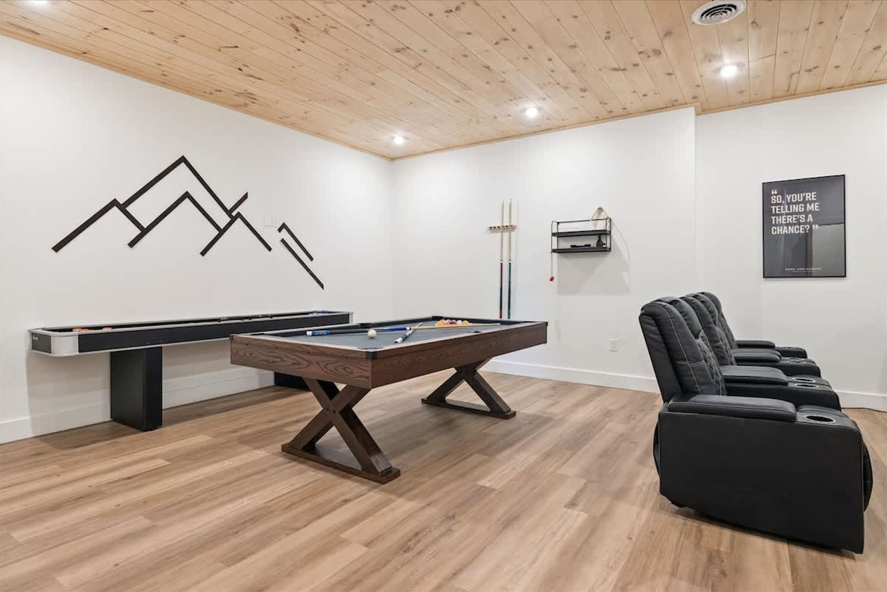 Une table de billard dans une pièce au plancher en bois, avec un canapé noir et un mur orné d’un motif géométrique.