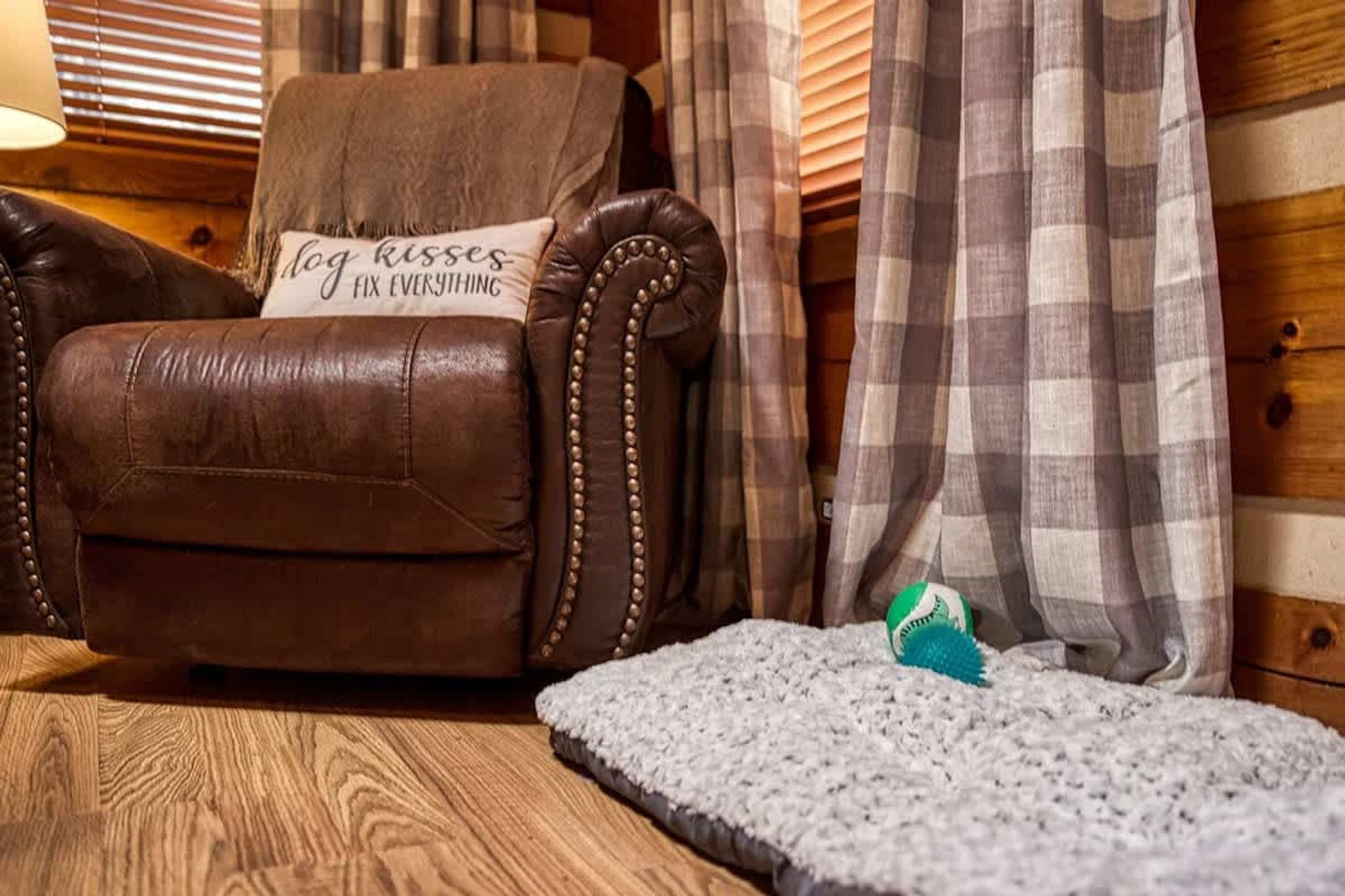 Uma poltrona de couro marrom com uma almofada com a frase 'dog kisses fix everything', uma cortina xadrez e uma pequena cama para animais de estimação com um brinquedo.