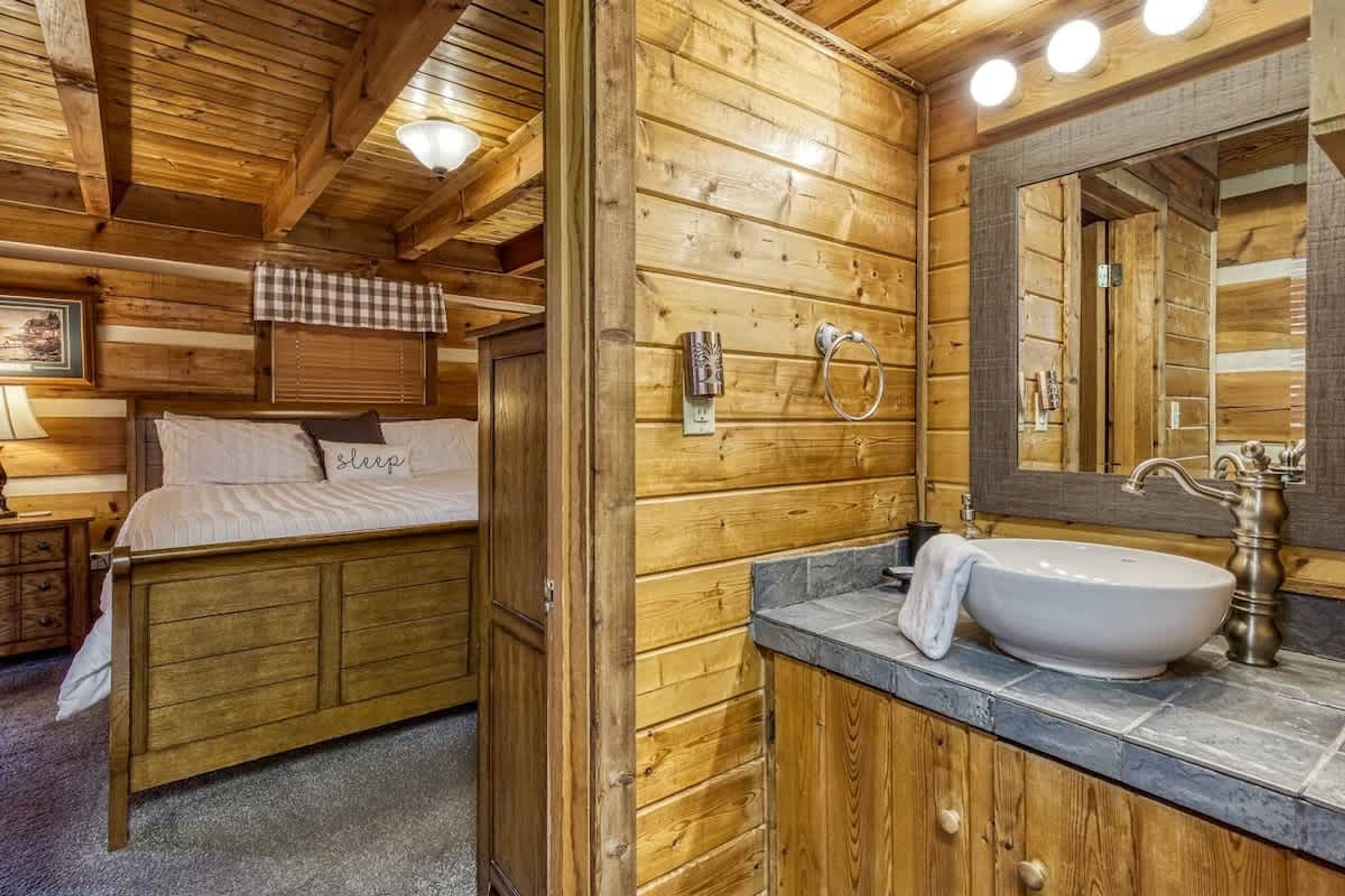 Ein Holzblockhaus Innenansicht mit einem Bett, einem Badezimmer mit Waschbecken und Spiegel sowie einem karierten Vorhang.