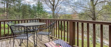 A wooden deck with a table and chairs, a bench with an American flag, and a view of trees and mountains.