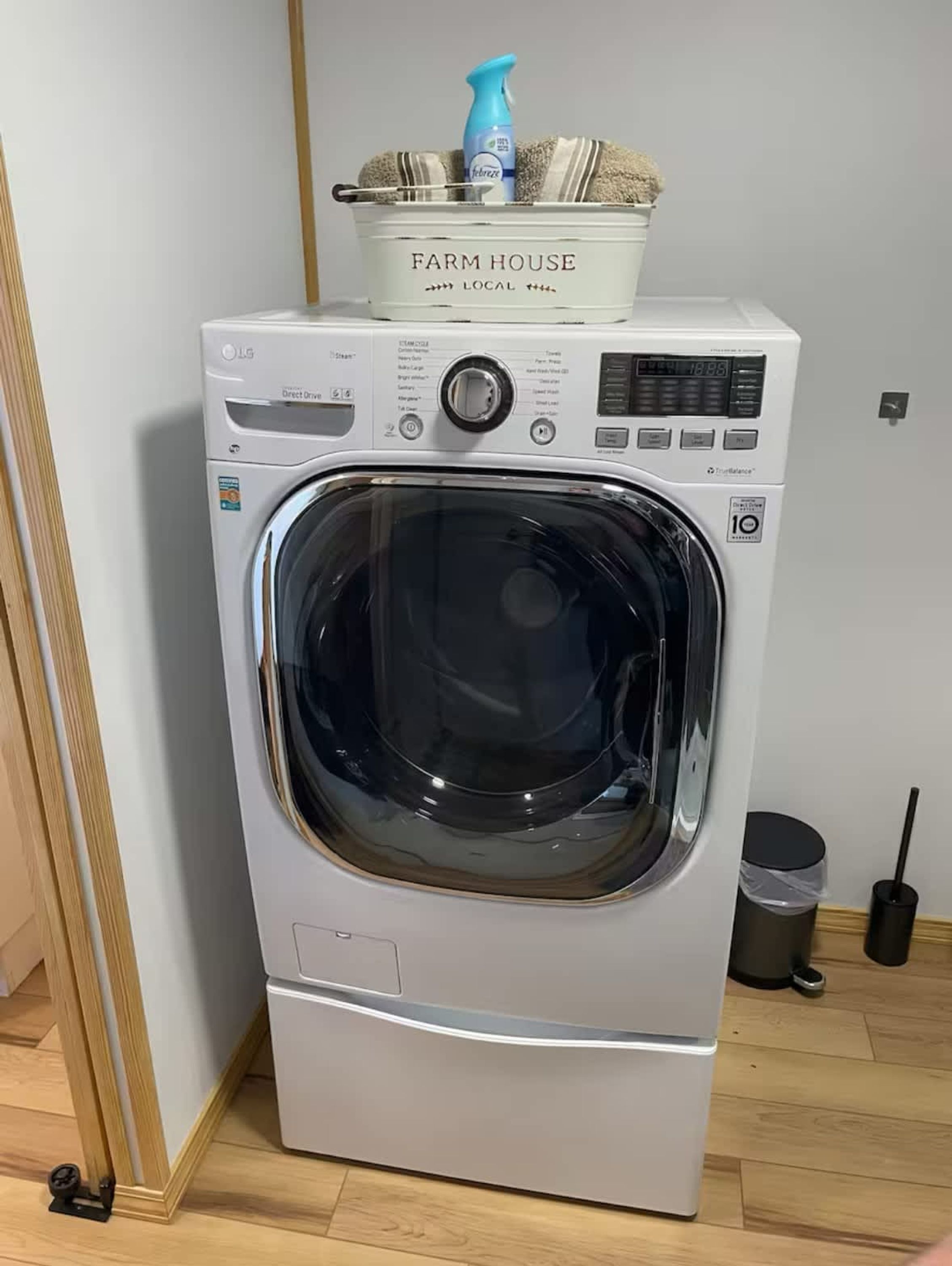 Une machine à laver LG à chargement frontal, surmontée d’un panier décoré dans le style « Farm House Local », dotée d’un écran numérique et accompagnée d’une poubelle placée à proximité.