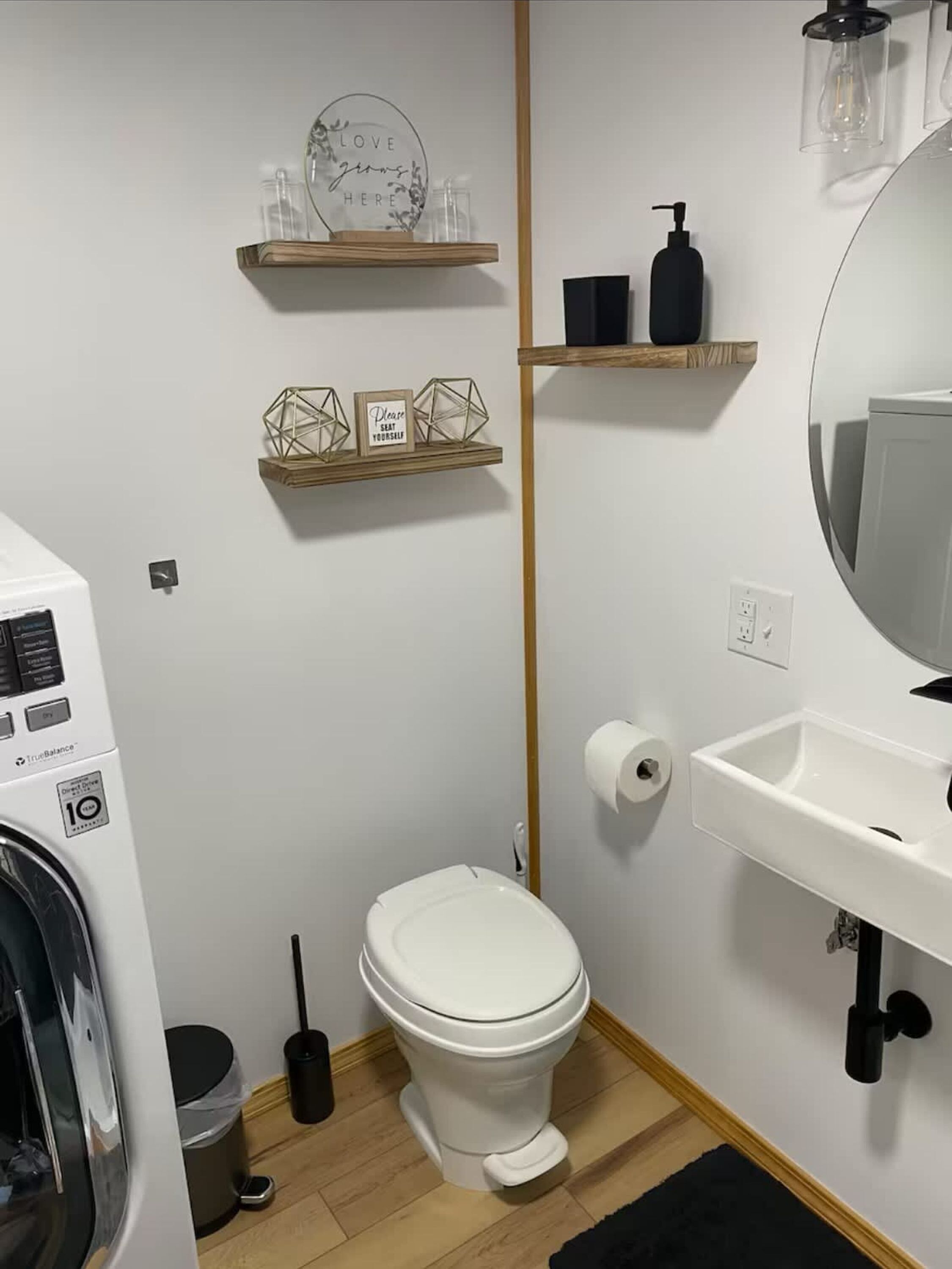 A small bathroom with a toilet, a sink, a mirror, a washing machine, and two shelves with decorative items.