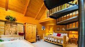 Interior de una cabaña de madera con una cama grande de madera, un armario de madera, una pared de piedra, una escalera de madera y un jacuzzi.