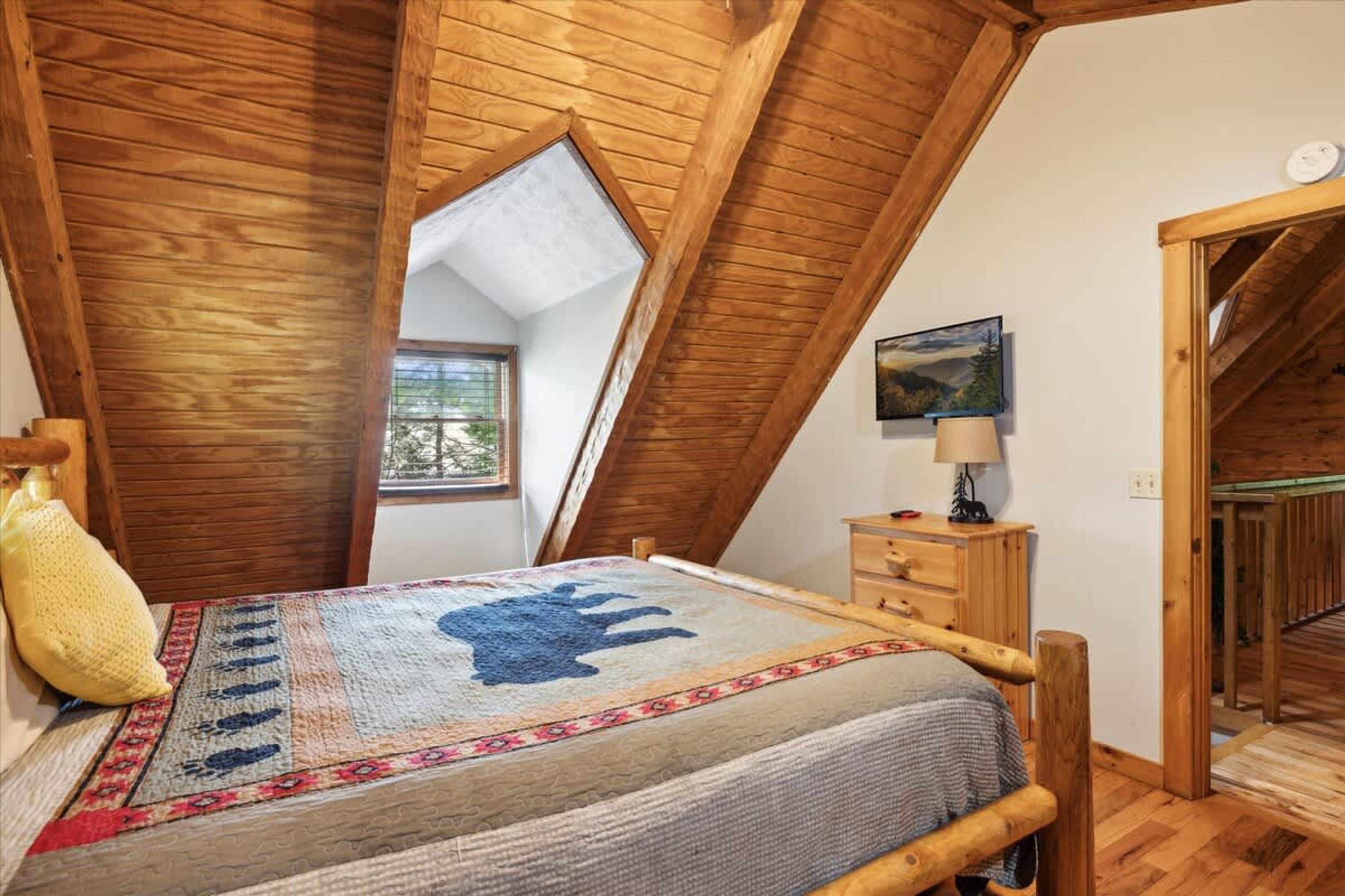 A bedroom with a sloped ceiling, a bed with a patterned bedspread, a bedside table with a lamp, and a window with a view of greenery.