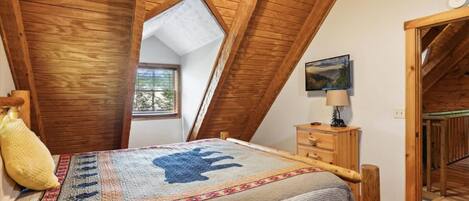 A bedroom with a sloped ceiling, a bed with a patterned bedspread, a bedside table with a lamp, and a window with a view of greenery.
