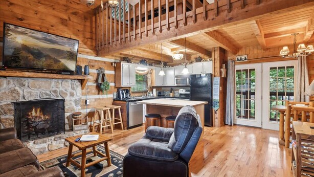 A log cabin interior with a stone fireplace, a leather sofa, a dining area, a kitchen, and a balcony with a view of the forest. - Cabin Fever by Beyond Expectations With Hot Tub (Gatlinburg)