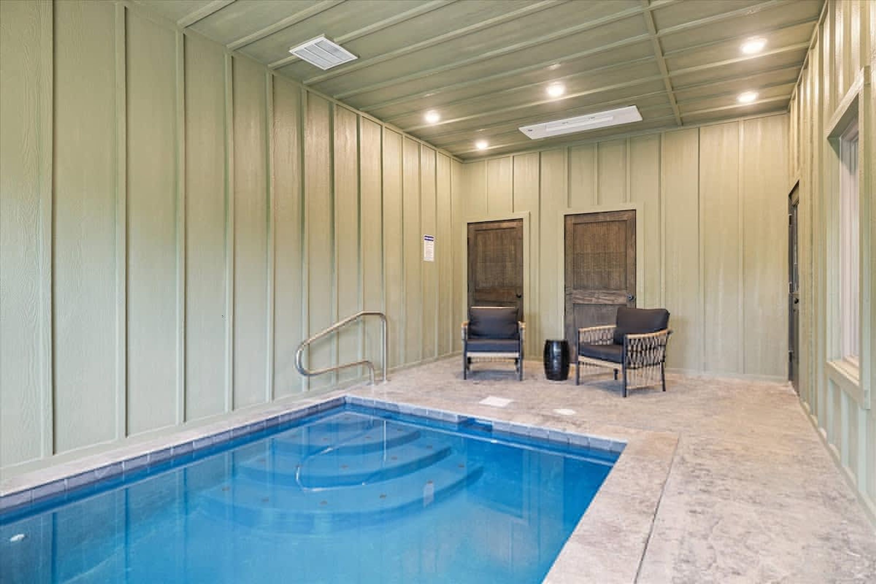 An indoor swimming pool with a tiled floor, two chairs, and a door.