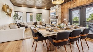A modern dining area with a wooden ceiling, a stone fireplace, and a large window with blinds. - Peak Pursuit by Beyond Expectations Mountain Home With Indoor Pool (Pigeon Forge)