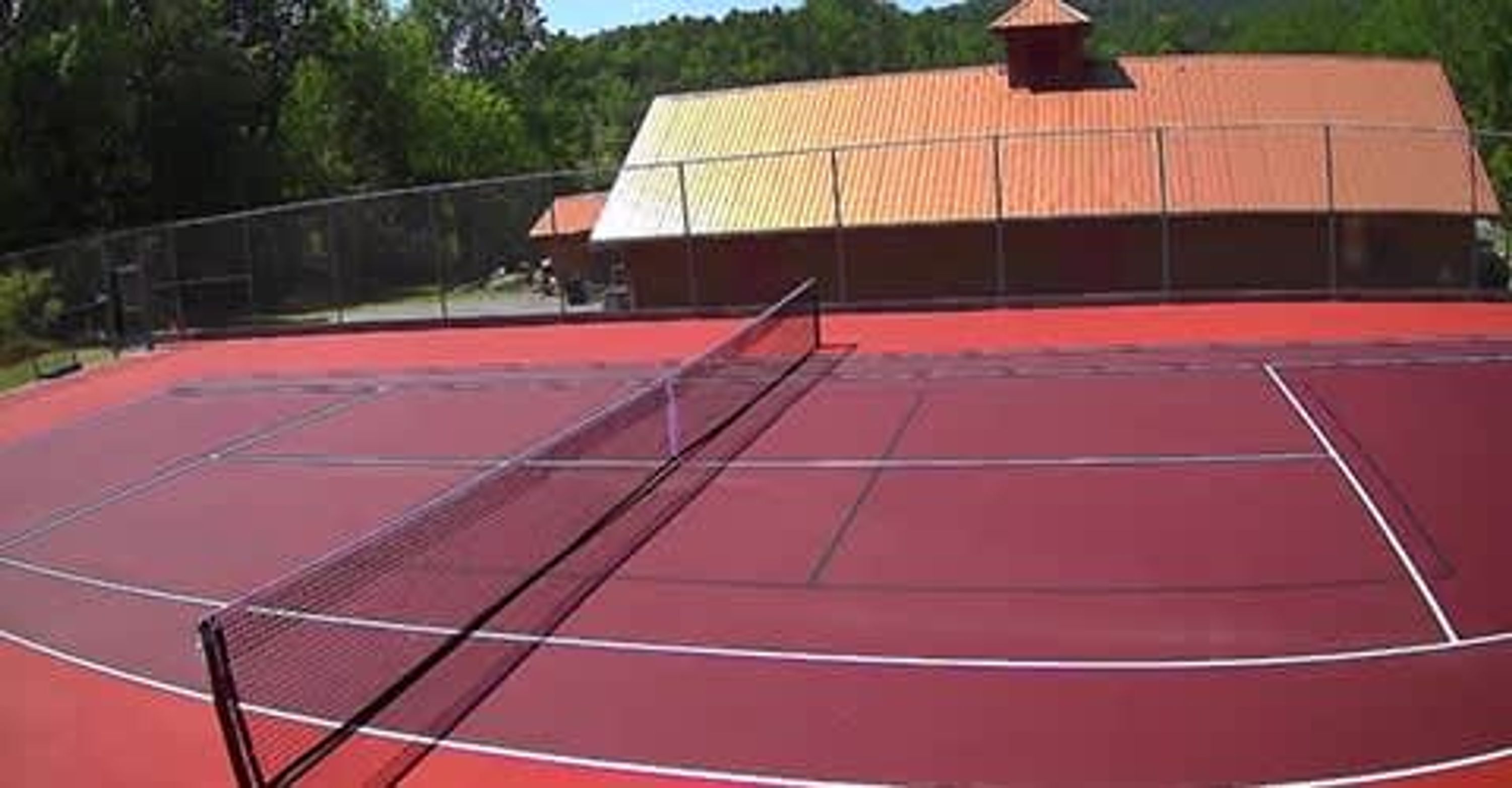 Een tennisbaan met een net, omringd door een hek en een gebouw met een rood dak.