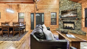 A log cabin interior with a stone fireplace, wooden dining table, and a cozy seating area. - Dotties Place Cabin by Beyond Expectations With Hot Tub (Gatlinburg)