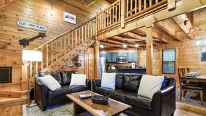 A wooden cabin interior with a living area featuring two leather sofas, a wooden coffee table, and a staircase leading to a kitchen area. - Dotties Place Cabin by Beyond Expectations With Hot Tub (Gatlinburg)