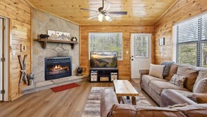 L’intérieur d’une cabane en rondins, avec une cheminée en pierre, un ventilateur de plafond, un canapé et un téléviseur.