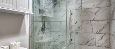 A glass-enclosed shower with marble tiles, a white cabinet above, and a shelf with toilet paper.