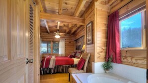 A wooden cabin interior with a bedroom featuring a bed, a red blanket, a chair, a window with curtains, and a bathroom with a bathtub. - Snuggle Up Cabin by Beyond Expectations With Hot Tub (Sevierville)