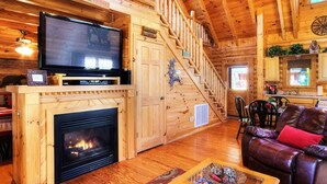 A cozy log cabin interior with a fireplace, a television, a leather sofa, and a staircase leading to the upper level. - Snuggle Up Cabin by Beyond Expectations With Hot Tub (Sevierville)