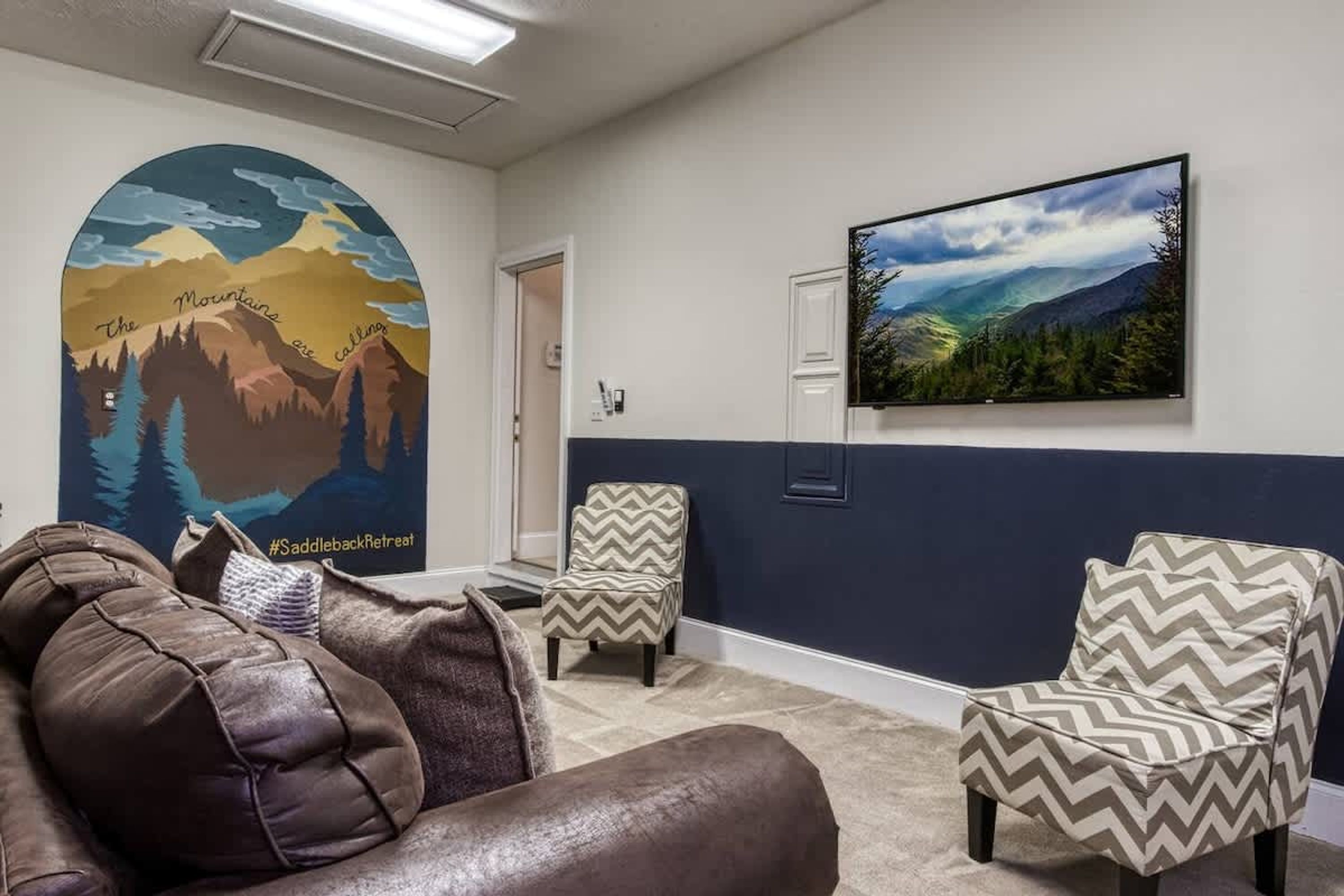 Una stanza con un affresco raffigurante montagne, una televisione, due poltrone a fantasia e un divano in pelle.