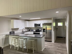 Una cocina moderna con armarios blancos, un refrigerador de acero inoxidable, un microondas y un horno empotrado. Hay tres taburetes blancos en la isla de la cocina.