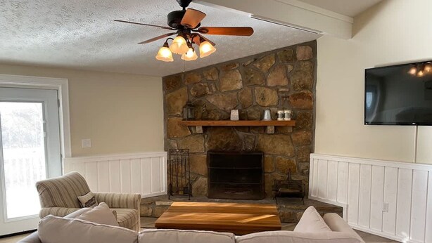Una sala de estar con chimenea de piedra, ventilador de techo, televisor de pantalla plana y un sofá seccional beige grande.
