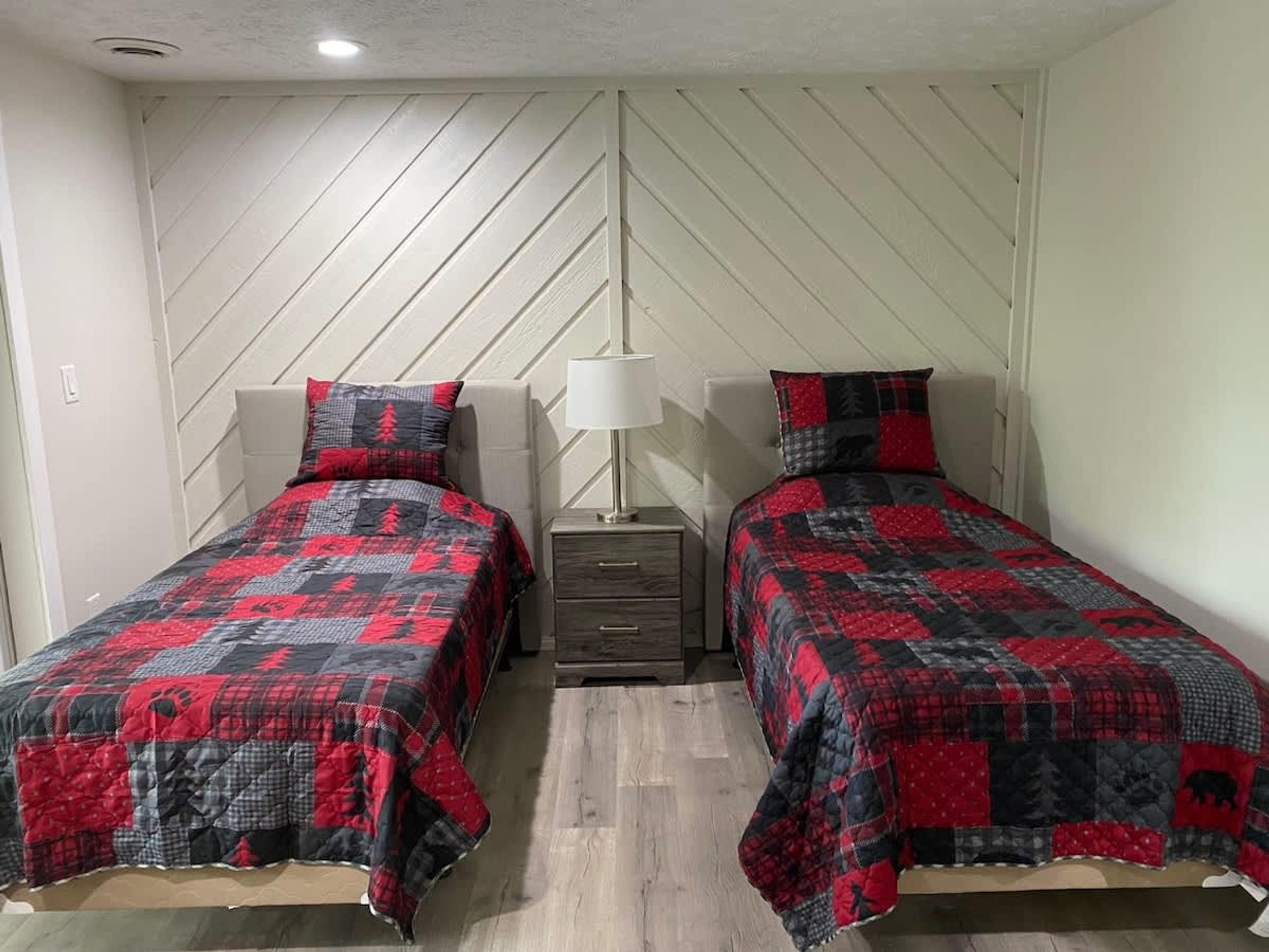 A room with two beds, each with a red and black quilt, a bedside table with a lamp, and a white wall with a diagonal pattern.