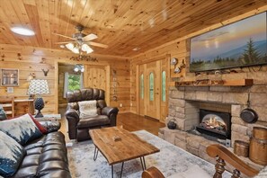 A log cabin interior with a fireplace, leather sofa, wooden coffee table, and a ceiling fan. - Chasing Cubs Cabin by Beyond Expectations (Sevierville)