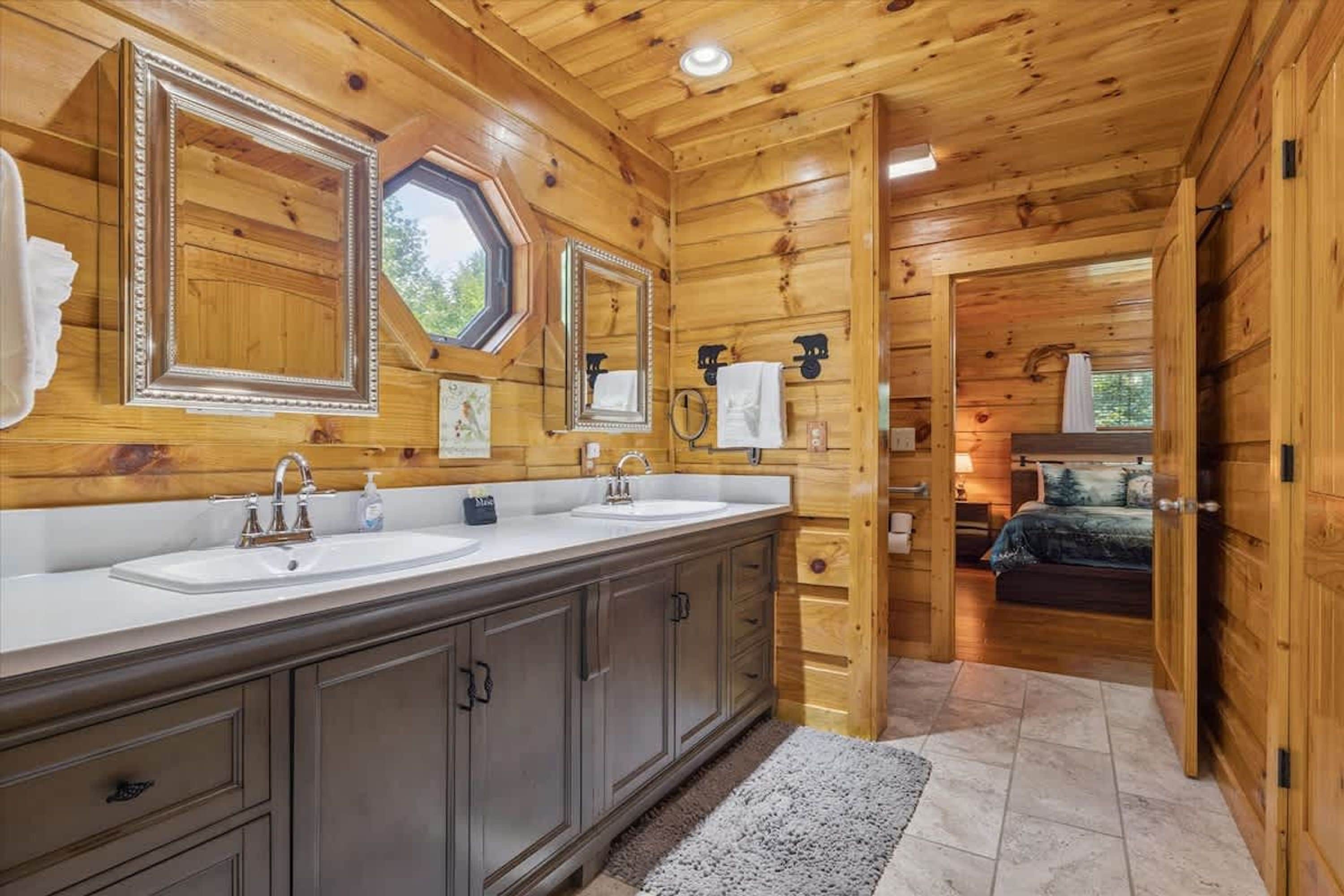 A wooden cabin interior with a double sink vanity, a mirror, and a bed visible through an open door.