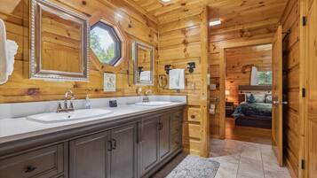 A wooden cabin interior with a double sink vanity, a mirror, and a bed visible through an open door.