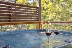 Un jacuzzi con dos vasos de vino tinto sobre una mesa, rodeado de listones de madera y follaje.