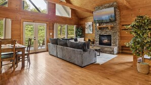 Ein Blockhaus-Innenraum mit Kamin aus Stein, Holzböden, einem grauen Sofa und großen Fenstern.
