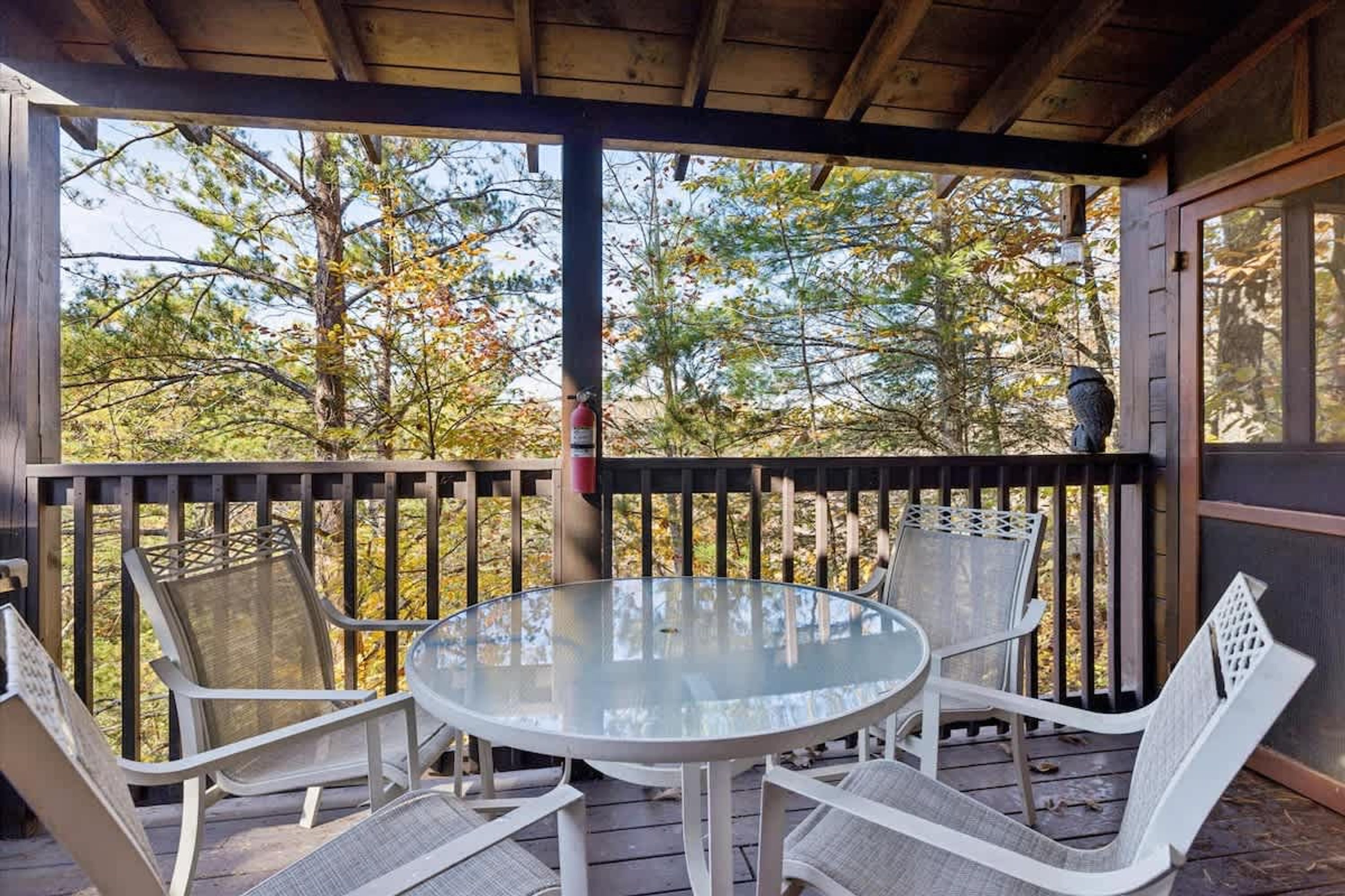 A covered outdoor seating area with a round table and chairs, a fire extinguisher, and a view of trees.