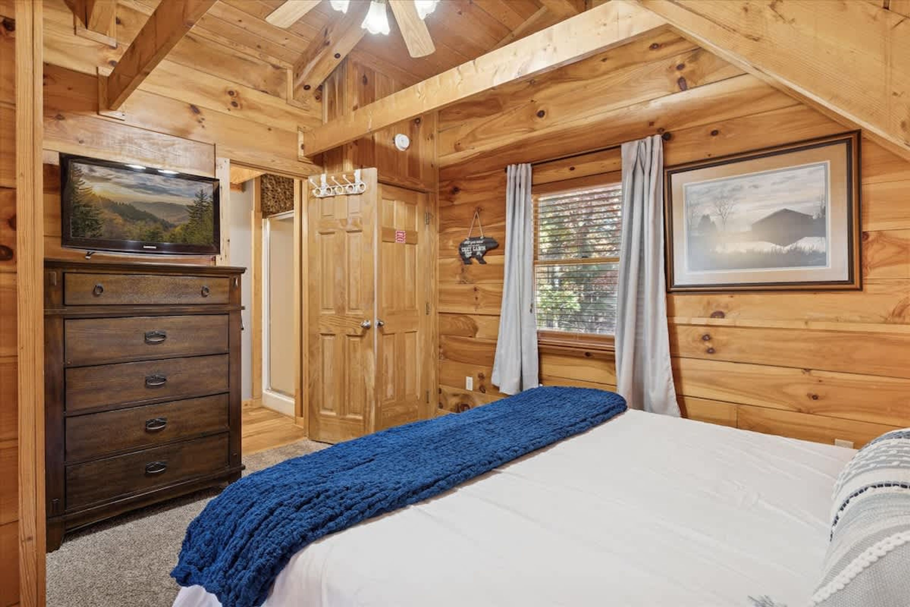 A wooden cabin bedroom with a bed, dresser, TV, and framed pictures.