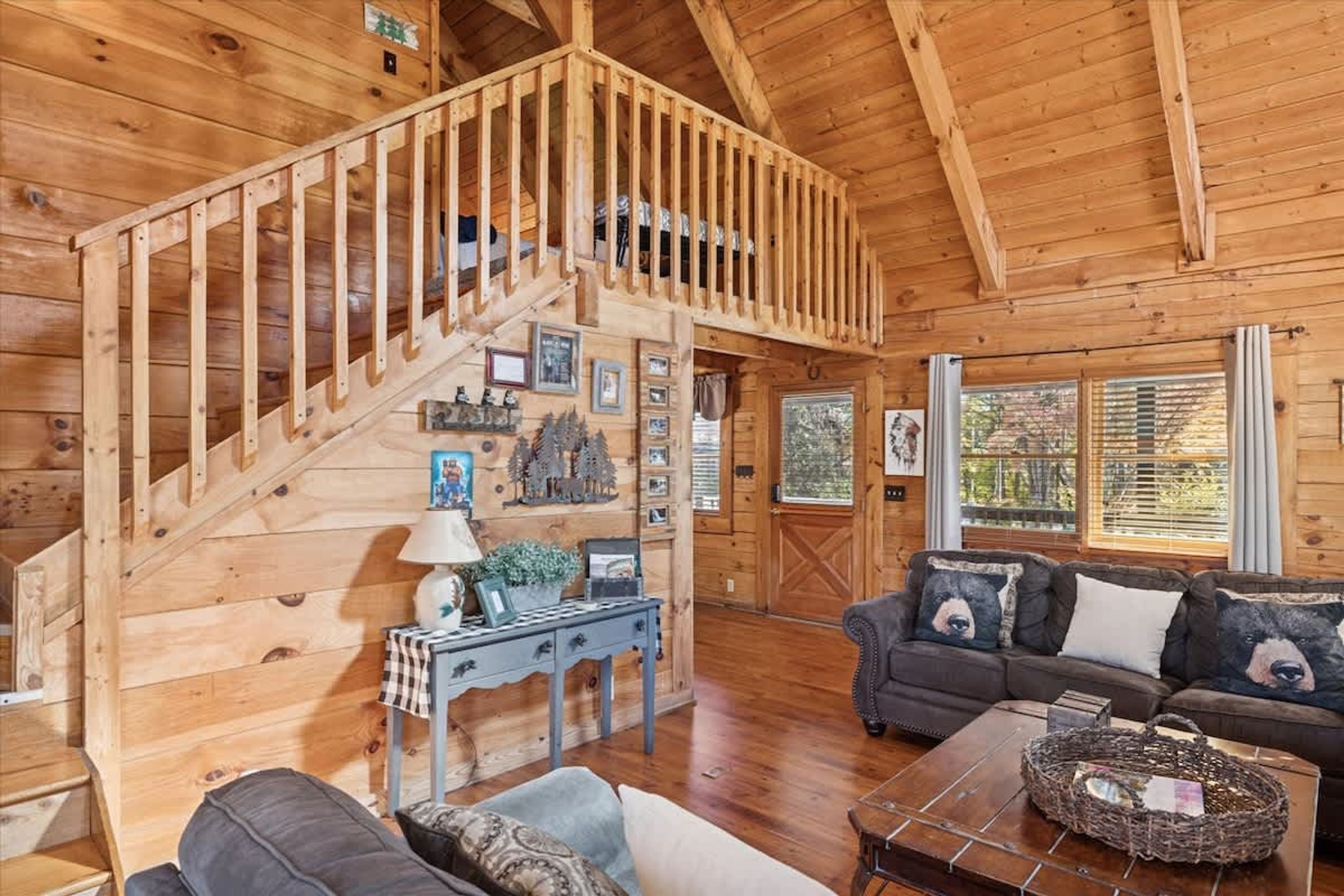 Een houten chalet van binnen met een trap, een bank met kussens met berenmotieven en een eettafel.