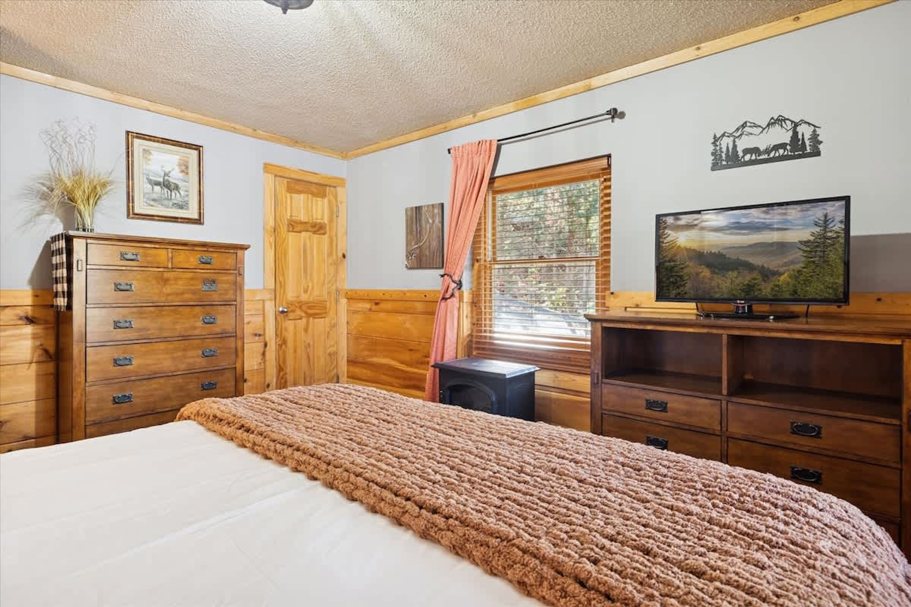 A bedroom with a bed, wooden dresser, television, and a window with blinds.