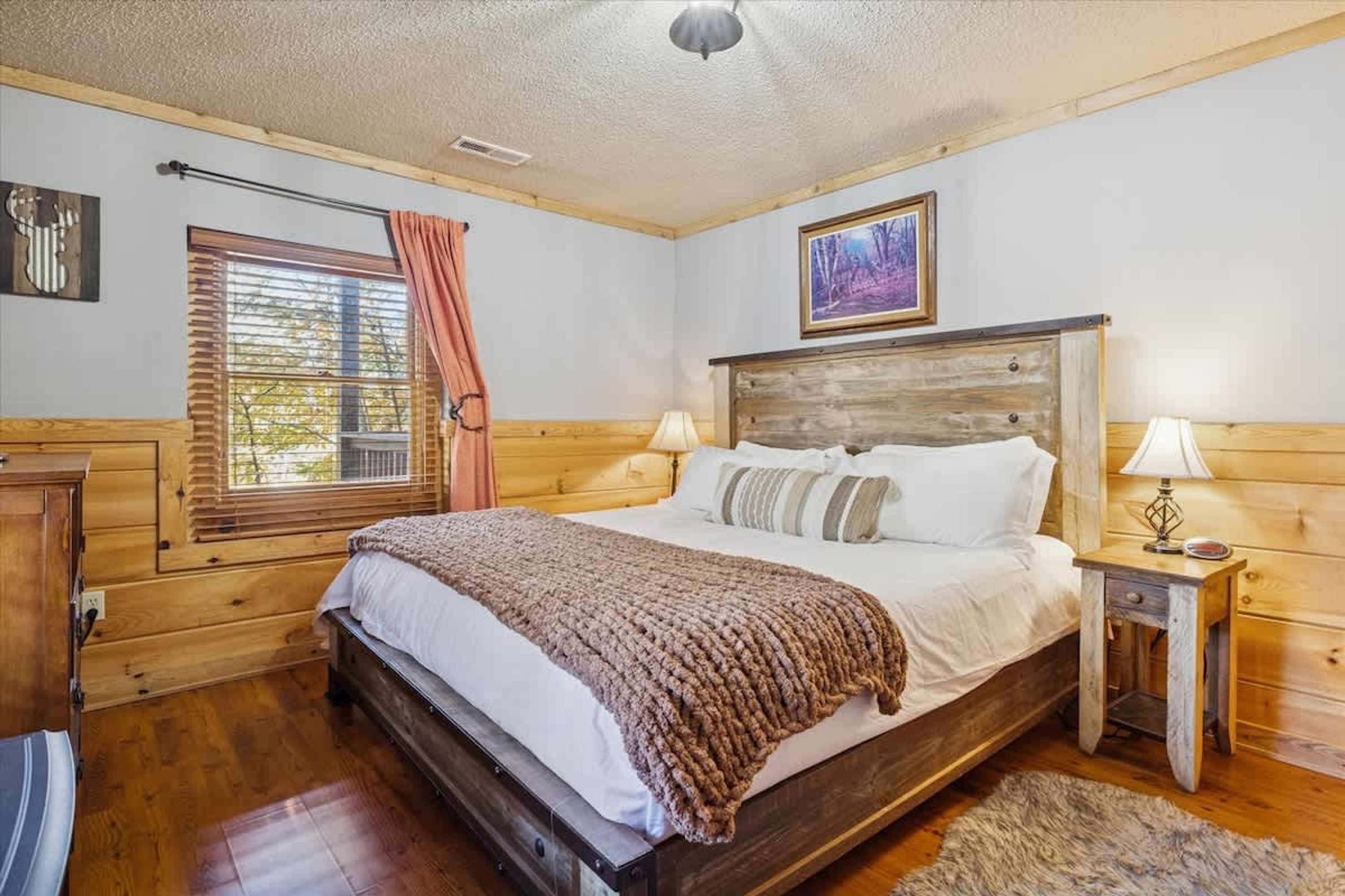 Camera da letto con struttura in legno, un letto, una finestra con persiane, una lampada e un quadro appeso al muro.