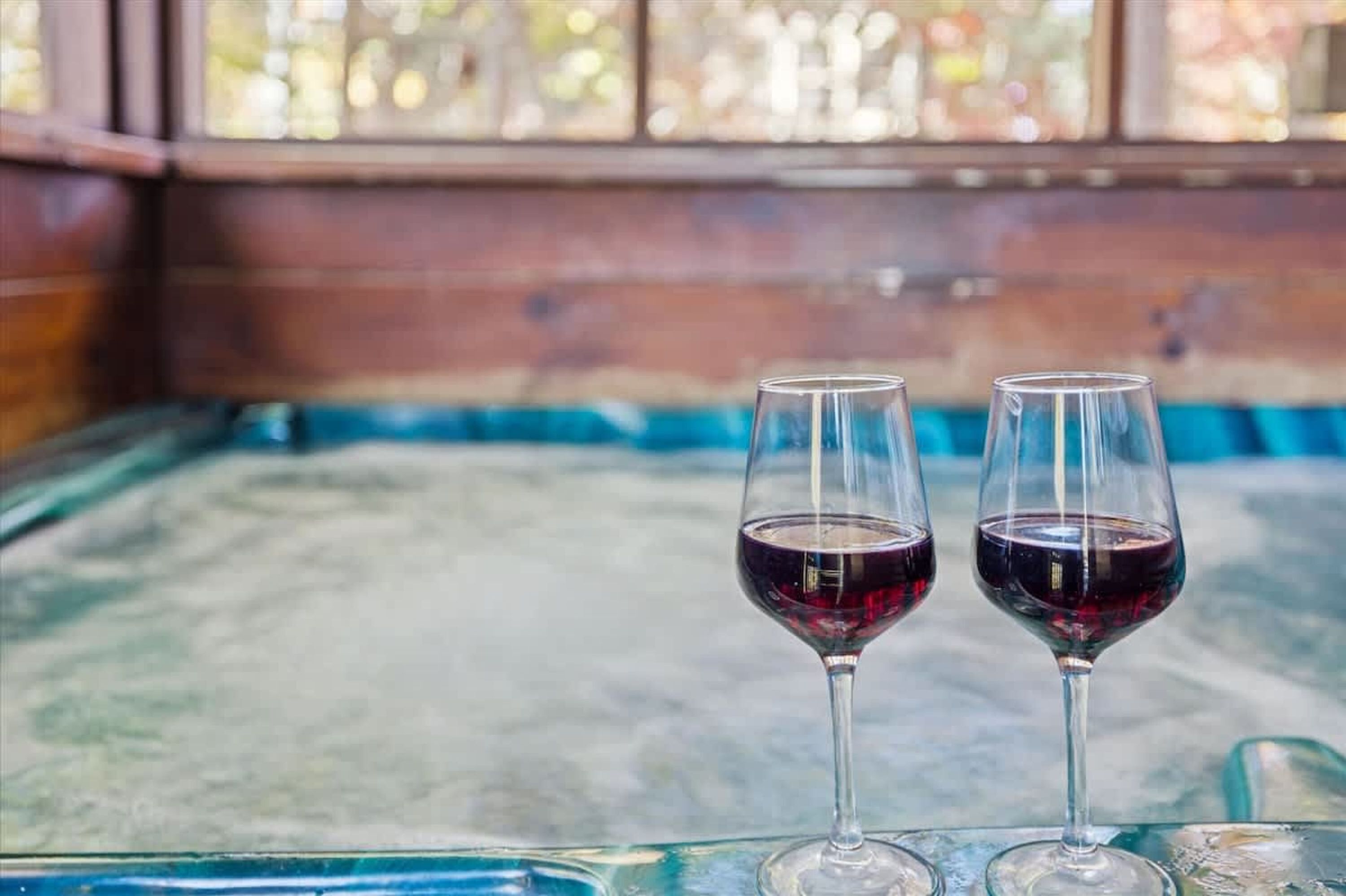 Two wine glasses on a glass table with a wooden bench and a window in the background.