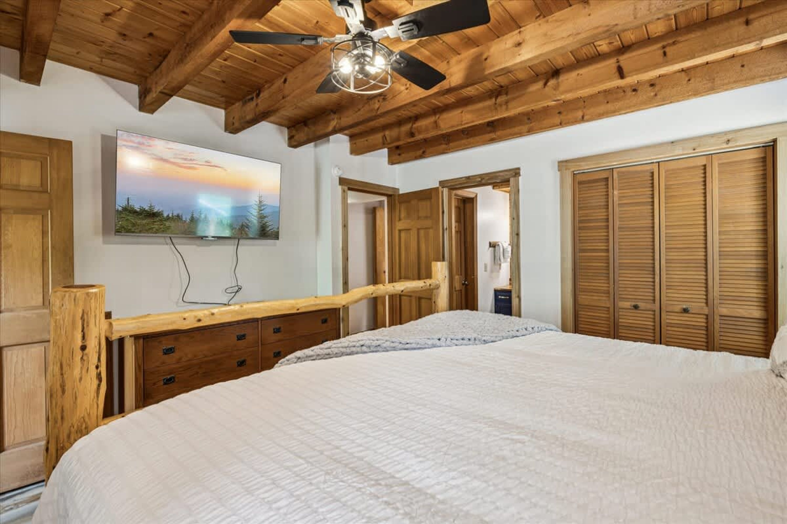 Un dormitorio con una cama grande, vigas de madera en el techo, un ventilador de techo, un televisor instalado y armarios de madera.