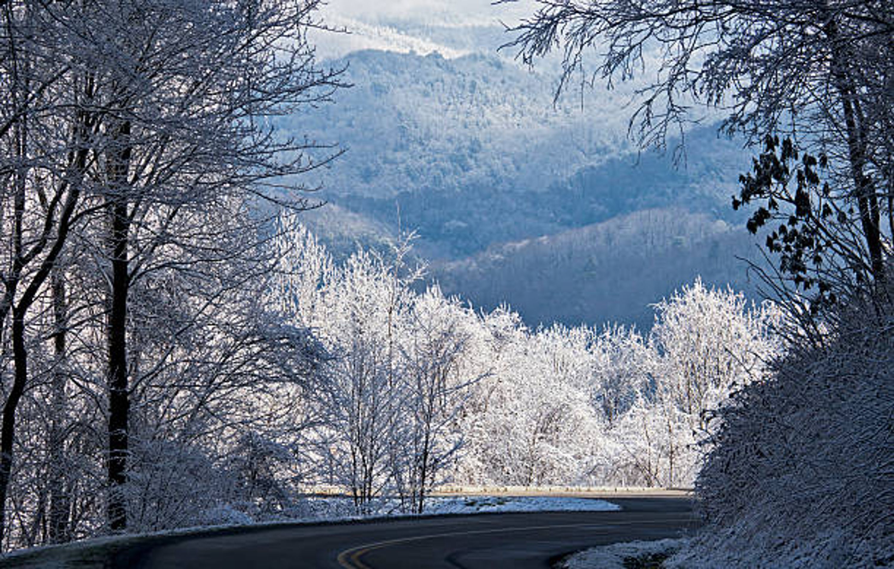 A snow-covered landscape with a winding road, trees, and distant mountains.