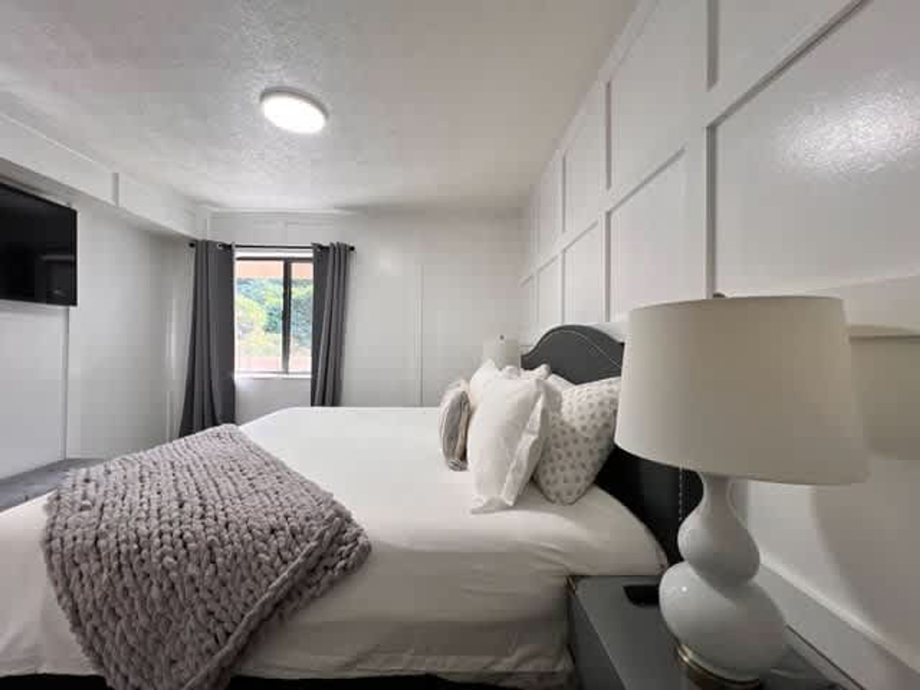 Habitación de hotel con cama, lámpara de noche, ventana con cortinas y televisor de pantalla plana montado en la pared.