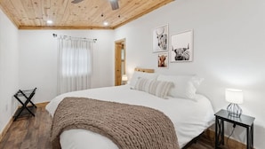 Un dormitorio con techo de madera, una cama con ropa de cama blanca, un edredón marrón, una mesita de noche con lámpara y dos cuadros enmarcados en la pared.