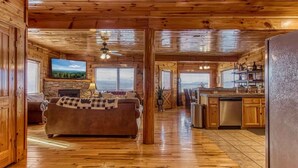 A log cabin interior with a living room, kitchen, and dining area, featuring wooden floors and a stone fireplace. - Majestic Overlook II by Beyond Expectations (Sevierville)