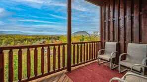 A wooden deck with a red mat, two chairs, and a view of houses and hills. - Majestic Overlook II by Beyond Expectations (Sevierville)