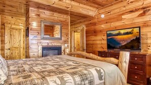 A log cabin room with a fireplace, a bed with a patterned bedspread, a mounted TV, and wooden walls. - Majestic Overlook II by Beyond Expectations (Sevierville)