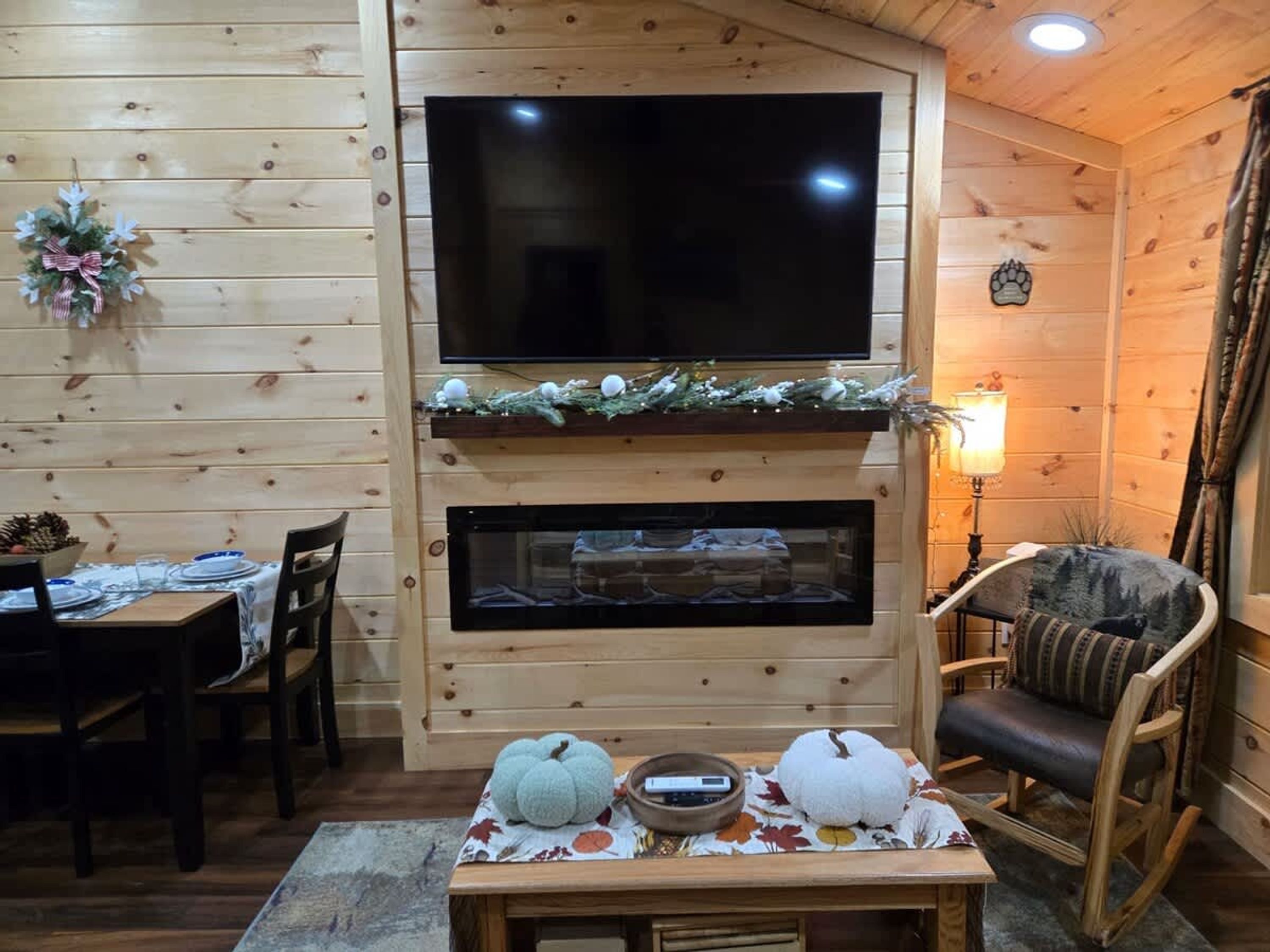 A cozy room with a TV mounted on a wooden wall, a fireplace below, a dining table with chairs, and a rocking chair.
