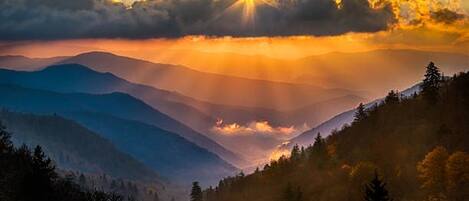 A mountainous landscape with a sunrise or sunset, trees, and a clear sky.