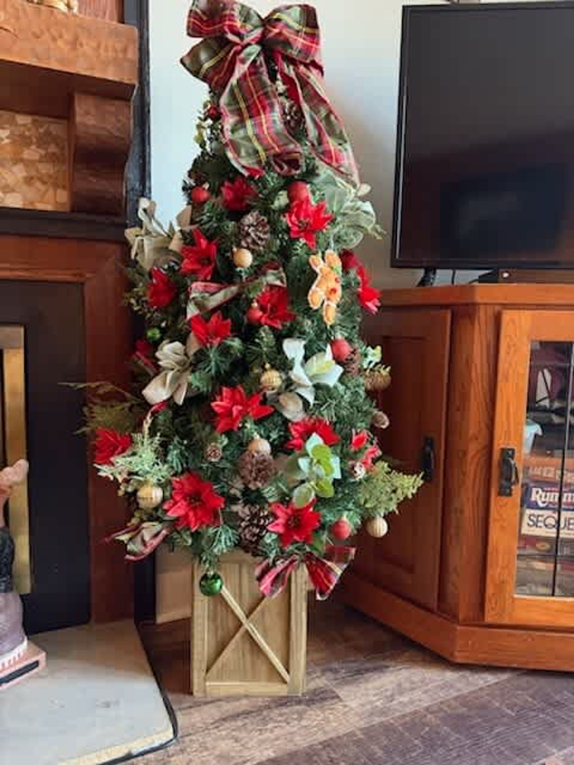 Un árbol de Navidad con adornos y un lazo, situado dentro de un mueble de madera con un televisor encima.
