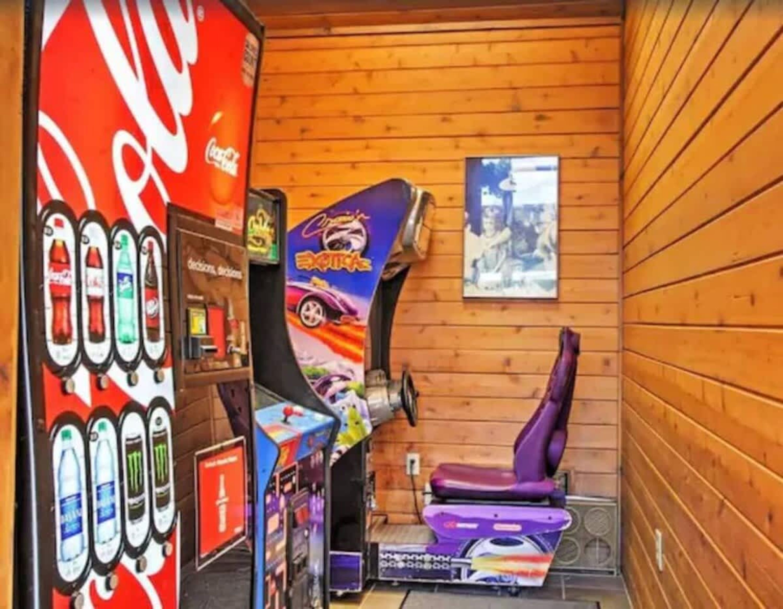 A room with wooden walls, a Coca-Cola vending machine, a poster of a racing game, and a purple gaming chair.