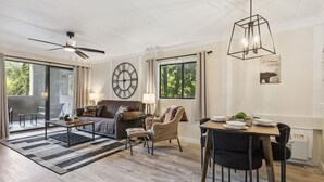 Amplia sala de estar con sofá, mesa de centro y mesa de comedor. Hay un ventilador de techo, un reloj y una ventana con cortinas.